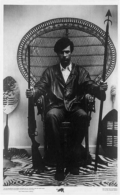 Huey Newton seated in rattan chair holding a rifle and spear