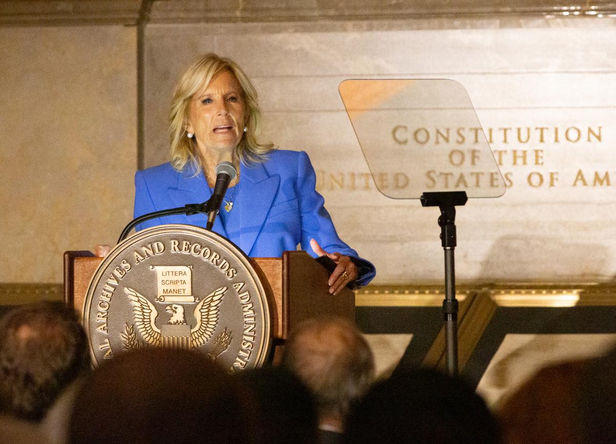 A white woman in a blue jacket gestures from behind a podium.