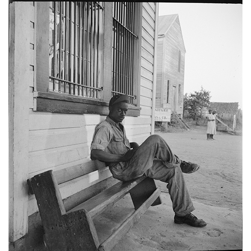 Black and white photo of a young black man sitting on a bench, staring at the camera, wearing work clothes.