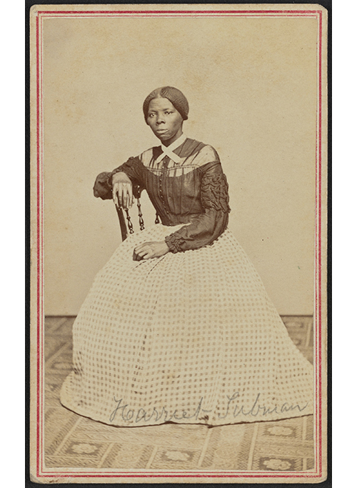 Photograph shows Harriet Tubman at midlife. She is seated, turned toward the left. One hand rests on the back of a wooden chair, another rests in her lap.