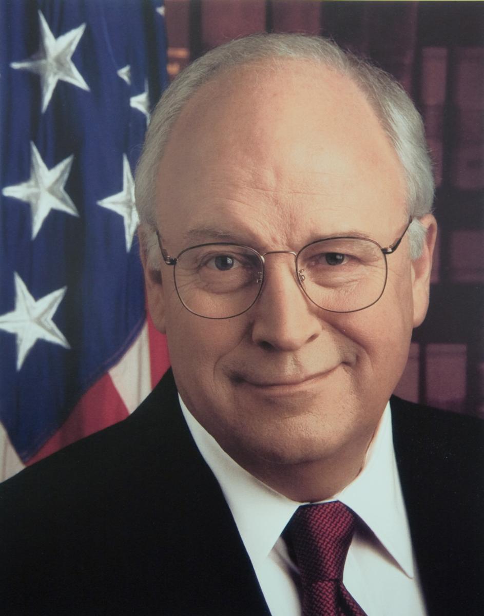 Vice President Dick Cheney smiling in a dark suit and red patterned tie, in front of an American flag and a bookcase.