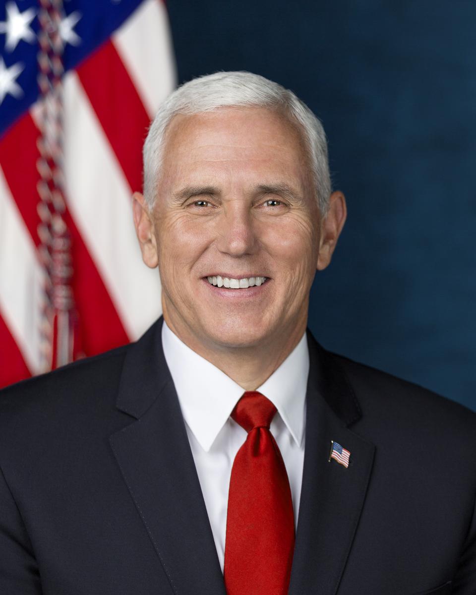 Vice President Mike Pence, smiling and wearing a dark suit, red tie, and American flag lapel pin, in front of an American flag.