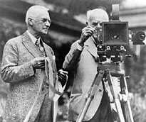 George Eastman holds the flexible 35mm film he invented, and Thomas Edison operates his movie camera