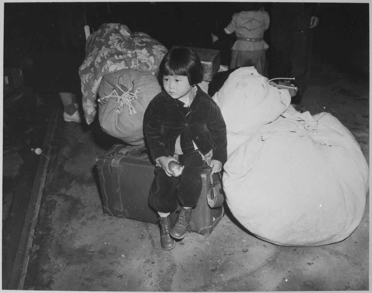 A young girl waits with the family baggage before leaving by bus for an assembly center
