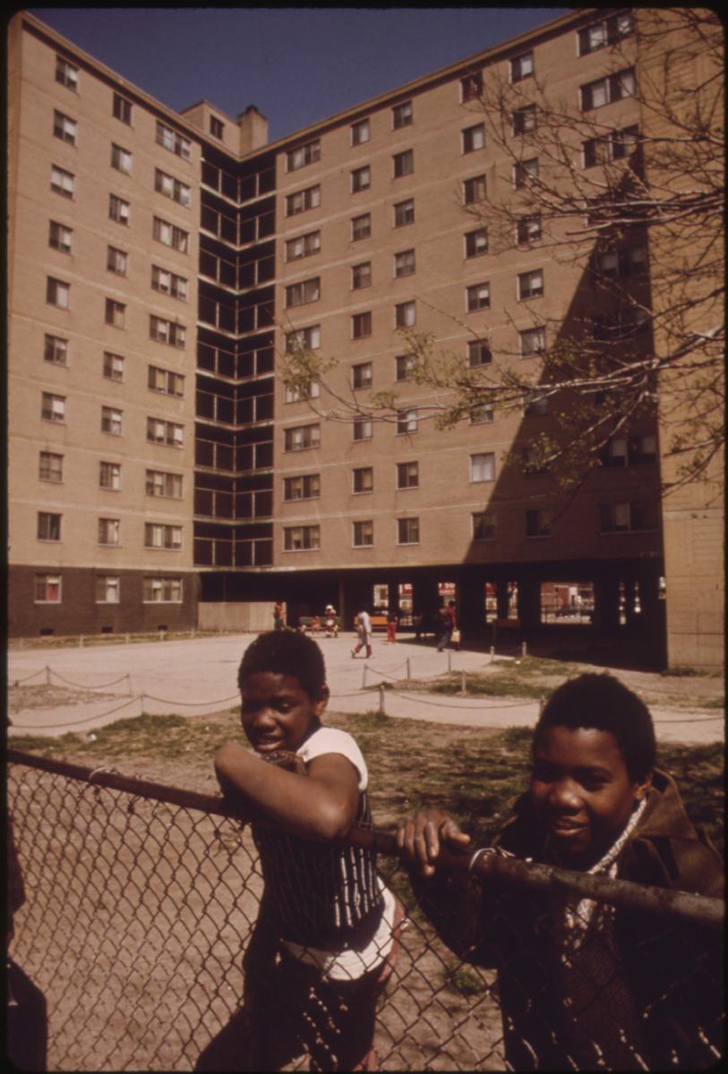 2 boys stand at fence with a highrise apartment building behind them