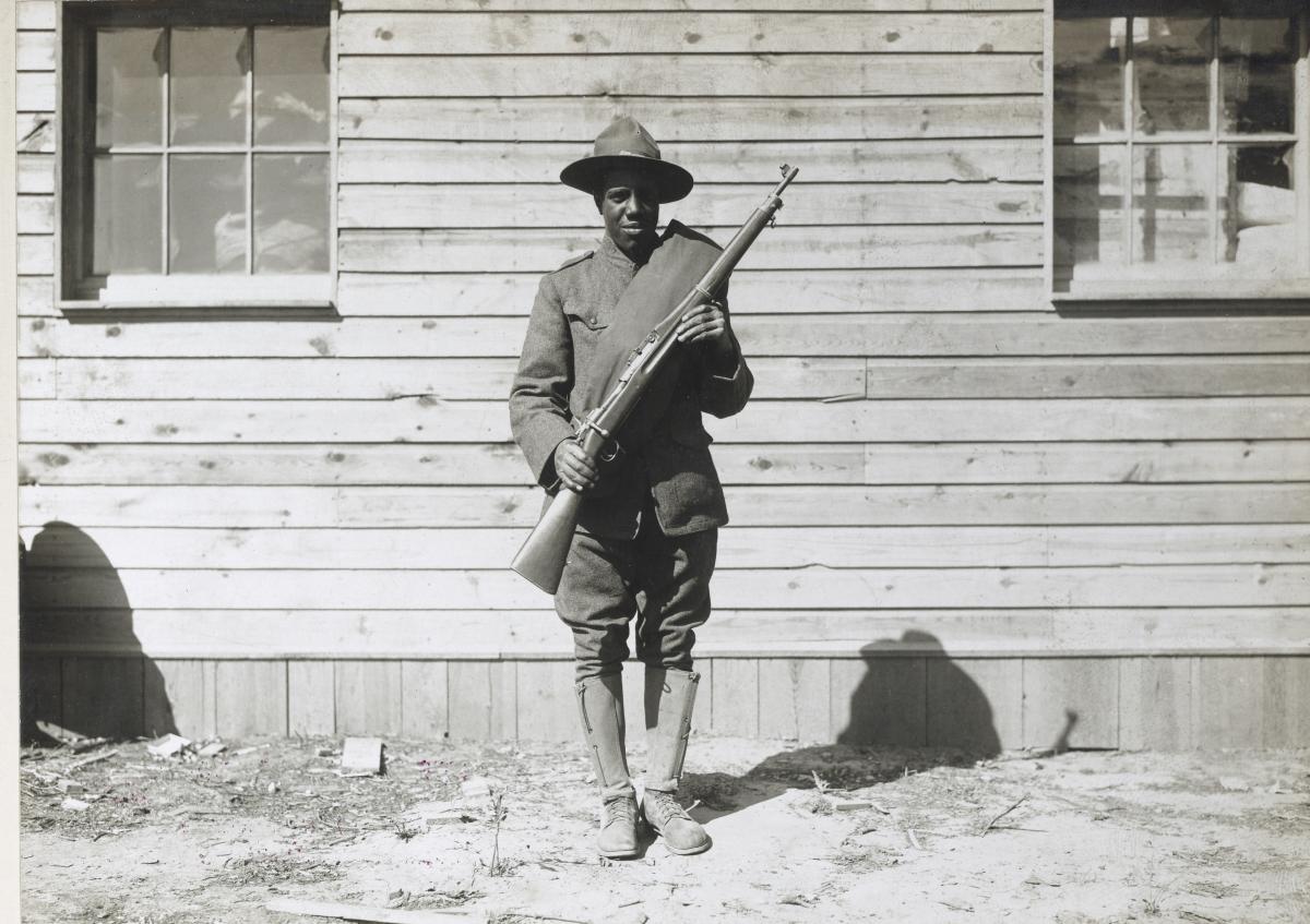 male soldier in uniform holding a rifle standing in front of a wooden building