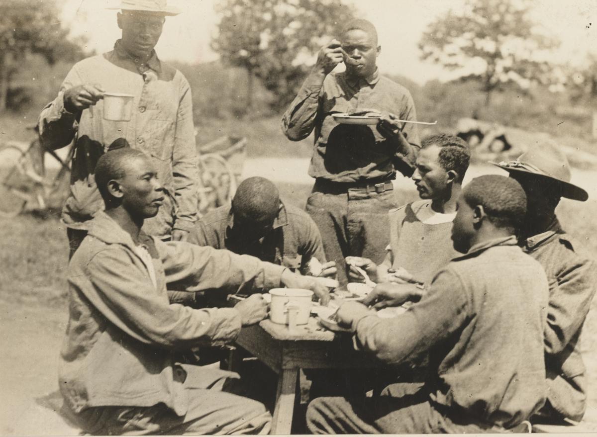 5 soldiers sitting at a table (2 standing) outdoors, eating out of skillets