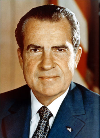 This is a photo of Richard Nixon behind his desk with the American flag to the right and leaning forward.
